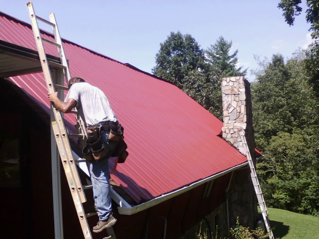 Roofer on a ladder beside a red metal roof and stone chimney — Soffit & Fascia Repair by Roofing Brothers in Sierra Madre